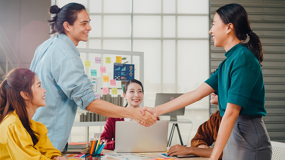 Multiracial group of young creative people in smart casual wear discussing business shaking hands together and smiling while standing in modern office. Partner cooperation, coworker teamwork concept.
